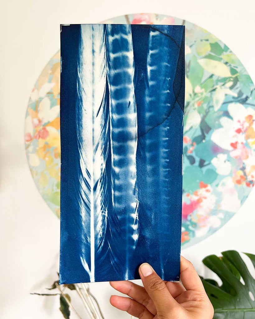Hand holding a blue feathered wall art with a floral background by artist Ingrid Sanchez.