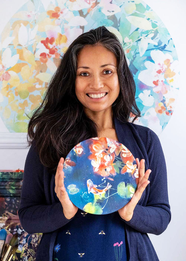 Ingrid Sanchez in her studio holding Mini Koi Pond, an original silk painting featuring a koi fish swimming amidst lotus leaves with cherry blossom reflections. The artwork, painted with dyes on silk and mounted on a 20 cm round wood panel, reflects her exploration of abstraction and nature. This photo highlights the painting’s scale and craftsmanship, offering a glimpse into the artist’s creative process.