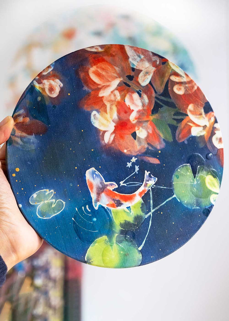 Mini Koi Pond, original silk painting by Ingrid Sanchez, featuring a serene koi fish swimming amidst green lotus leaves with delicate cherry blossom reflections above. Painted with dyes on silk and mounted on a 20 cm round wood panel (1.8 cm thick), this unique artwork blends abstraction with nature. Perfect for small spaces, this ready-to-hang piece adds calm and beauty to any home or office décor.